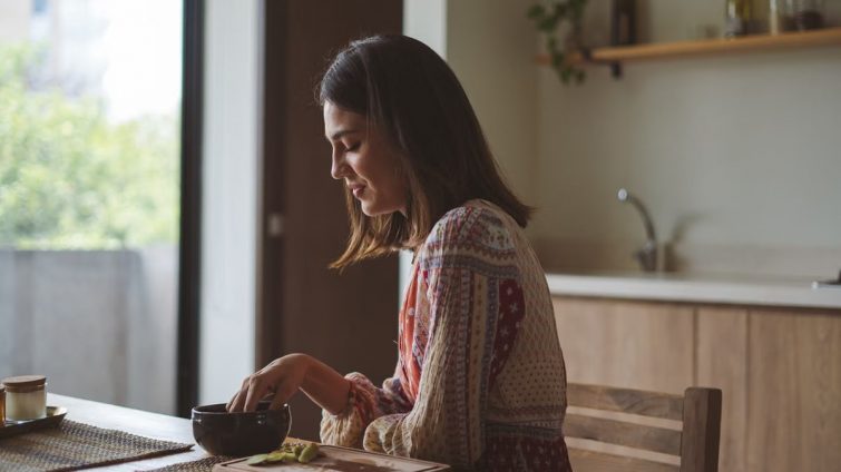 Woman in patterned dress enjoying breakfast at home near a sunlit window.