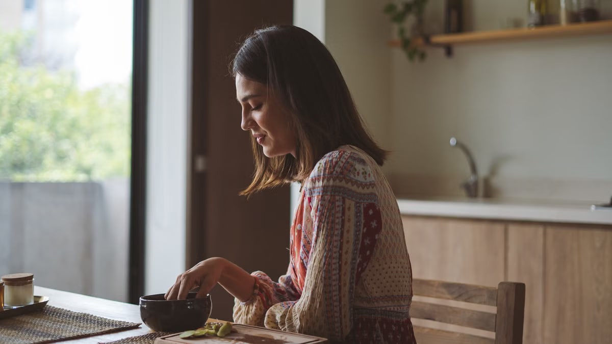 Woman in patterned dress enjoying breakfast at home near a sunlit window.
